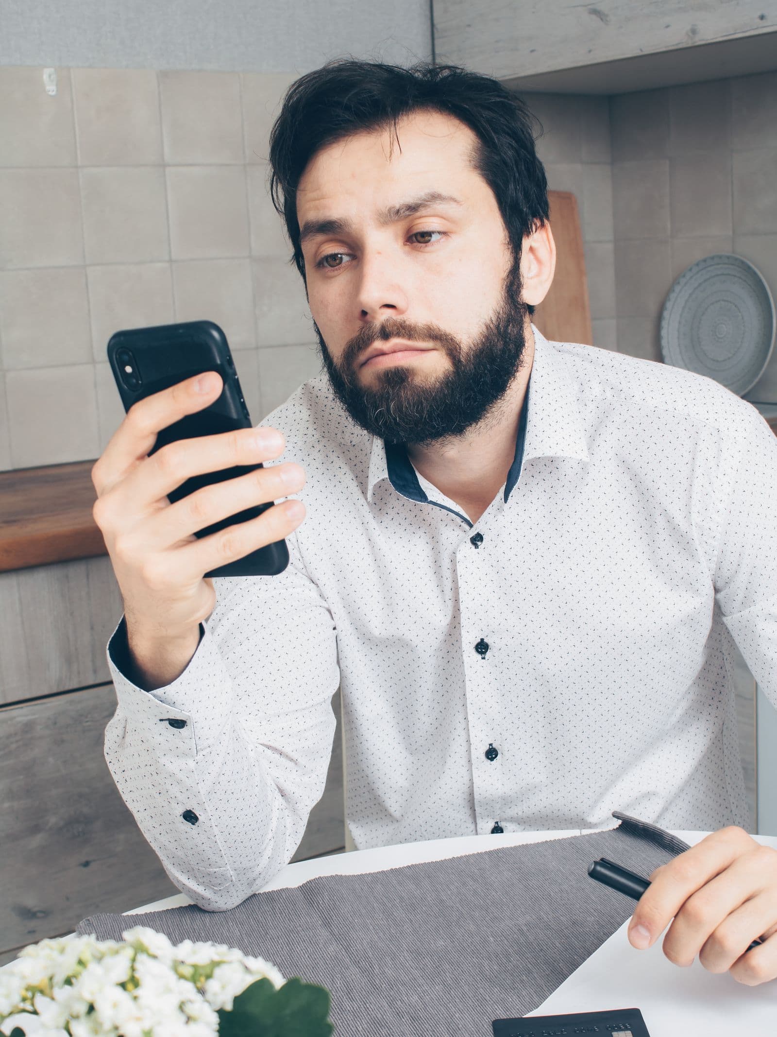 Homem olhando desconfiado para a fatura no celular, com cartão de crédito sobre a mesa